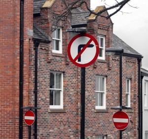 From Crossgate. Three vertical road signs on a typically English brick building background. Black drainpipes visually merge with black sign posts.