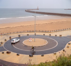 Sunderland, England - August 5, 2012: After a storm, the light conditions change the normal colour saturation of the scene: the sand appears almost as pink as the dress of one of the ladies in the centre of the roundabout. A car and a cyclist (with the usual ugly but useful yellow high-visibility vest) also arrive onto the scene immediately after the rain clears. The Roker pier is visible in the background. Scan from 120 Portra 400