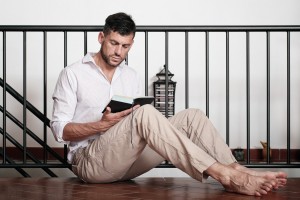 Man sits on the tiled floor, leaning his back onto the staircase's handrail, and reads a novel. Shallow soft focus. CD tower and small furniture in the background
